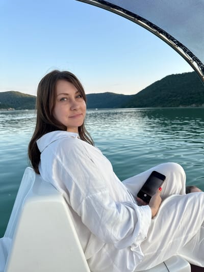 A woman with long, brown hair and blue eyes is captured in a vertical, outdoor portrait. She is wearing a white, oversized shirt and is seated on a white boat, holding a black smartphone in her right hand. The phone screen is dark, and the camera is facing the woman. Her fingernails are painted red. She is looking towards the camera with a slight smile.
The boat is on a body of water, likely a lake or reservoir, with calm, rippling green water. In the background, a lush green hillside or mountain range is visible, with a clear blue sky above. The image appears to be taken during the daytime, possibly late afternoon or early evening, given the soft lighting.
The setting suggests a leisurely activity, such as a boat trip or cruise, in a natural environment. The overall mood is relaxed and serene. The location context provided is Abrau-Dyurso, Russia, which is known for its wineries and lakes. There is no visible text in the image.