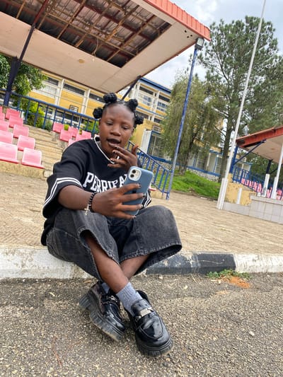 A young Black woman with dark skin and dark braided hair styled into four buns is sitting on a concrete curb outside. She is wearing a black long-sleeved shirt with white stripes on the sleeves and the word "PERFECT" printed across the chest. Her pants are black denim with a slit up the side of each leg, and she is wearing dark, chunky platform loafers with grey socks. She has a beaded bracelet with a cross charm on her left wrist and appears to be looking at and touching her lips while holding a blue smartphone in her right hand.

The background shows a stadium-like structure with pink seats visible on tiered bleachers. Behind the bleachers, a light yellow building with multiple windows is visible, with trees and foliage in front of it. The sky is overcast. The area appears to be an outdoor public space, possibly a school or sports facility, in the city of Zawan, Nigeria.