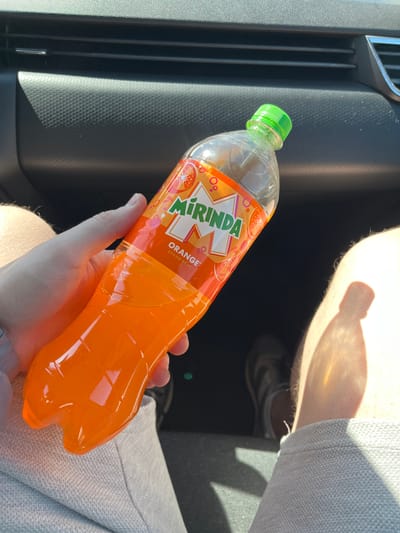 A person is holding a bottle of Mirinda orange soda in a car. The bottle is mostly full, with an orange liquid inside. The car interior has black plastic trim near the soda bottle and light gray fabric seating and possibly clothing visible around the hand holding the soda. The background features the interior of the car and a hint of a second person's leg and foot. There is no information to determine if this is Гудагайскі сельскі Савет, Belarus.