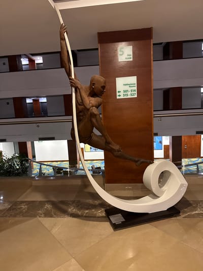 The image shows a sculpture of a gymnast holding a white curved bar on a brown tile floor inside a building, likely a hotel, in Querétaro, Mexico. The sculpture is positioned near a brown wooden wall with a sign indicating "5 PISO FLOOR" and another sign directing towards rooms "501-514" and "515-527." The building interior features a multi-story atrium with windows. The lighting is indoor, possibly artificial.