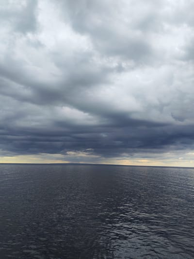 A wide, still body of water, likely a lake or a calm sea, occupies the majority of the frame. The water is dark and appears rippled, reflecting the overcast sky. The horizon line is sharp and divides the water from the sky, with a thin strip of land visible in the far distance. The sky is dominated by heavy, dark gray clouds, characteristic of stormy or unsettled weather. There are a few brighter patches of sky visible on the horizon, suggesting a break in the clouds or the last light of day. The overall scene is atmospheric and somewhat somber, with a sense of vastness and natural power. The location is the city of Shcheleiki, Russia.