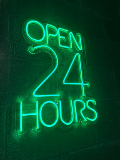 A neon sign glows green against a textured, dark-green brick wall. The words "OPEN 24 HOURS" are spelled out in neon tubing, arranged diagonally. "OPEN" is on the top left, "24" in the middle, and "HOURS" on the lower right. The bright green light highlights the brick surface, creating a stark contrast between the glowing sign and the dark background.