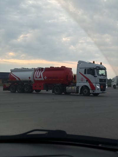 A white and red tanker truck, branded with the Lukoil logo, is parked on a paved surface under a cloudy sky. The truck's trailer is predominantly red with a white section near the front, featuring the Lukoil emblem. Text on the side of the trailer reads "ОГНЕОПАСНО," which translates to "Flammable." The truck cab is white with red accents and has a license plate visible. In the background, a few other trucks are parked and a building with a dark roof is visible. The scene appears to be an outdoor lot, possibly a truck stop or depot, located in Усть-Сыны, Russia.
