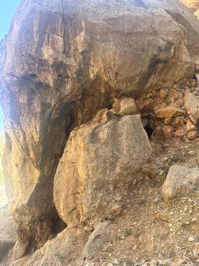 This media file depicts the underside of a rocky overhang, likely a cave entrance or a rock shelter. The scene is dominated by rough, textured rock formations in varying shades of brown, gray, and beige, with some areas showing lighter, almost white mineral deposits. The lighting suggests it is daytime, with sunlight casting shadows and highlighting the geological features. The rough, uneven surface of the rocks indicates a natural, outdoor environment, likely a mountainous or rugged terrain.

A small, dark opening, suggesting the entrance to a cave or a deep crevice, is visible in the upper-center portion of the image. Surrounding this opening are loose rocks and gravel, indicative of a natural accumulation of debris. The textures of the rocks range from large, weathered boulders to smaller, more fragmented pieces. Some parts of the rock surfaces exhibit darker, almost charcoal-like streaks, possibly from water erosion or mineral staining.

The perspective is from below, looking upwards at the rock ceiling, with a sliver of bright sky visible at the bottom and a hint of vegetation peeking through the rocks at the far right. The overall impression is of a raw, natural landscape. There is no visible text, people, or specific objects that would indicate a particular event or activity, other than the implied exploration of a natural geological formation. The lack of human presence and the ruggedness of the terrain contribute to a sense of wilderness and untouched nature. The location context of Ardales, Spain, suggests a Mediterranean or sub-tropical environment, which could explain the type of rock formations and the clear sky.