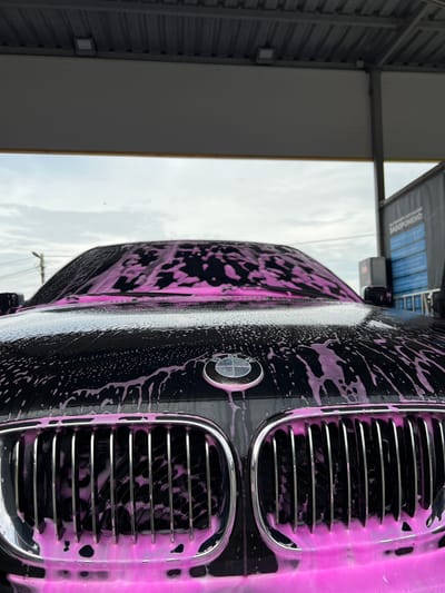 A low-angle, head-on perspective captures the front of a black BMW automobile, identified by its visible emblem and distinctive kidney grille, in the process of being washed at a self-service car wash facility in Velyki Hai, Ukraine. The entire visible front surface of the car, including the hood, windshield, and chrome grille slats, is thoroughly covered with a dense, bright pink cleaning foam. The foam is actively dripping and flowing, creating distinct rivulets and clinging in numerous small bubbles across the vehicle's dark finish. Overhead, a corrugated metal roof structure is visible, with a single white rectangular light fixture mounted underneath. The background reveals an overcast sky. To the right of the vehicle, a tall dark sign board, typical of car wash instructions, prominently displays the Ukrainian word "ЗАБОРОНЕНО:" (Forbidden:) followed by additional blue graphic elements and obscured text, confirming the geographical region. A utility pole with associated wiring is faintly visible on the left side of the horizon. No individuals are present within the frame.