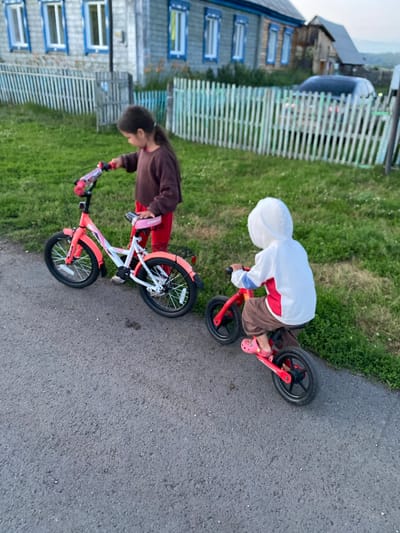 In Maloarslangulovo, Russia, two young children are engaged in outdoor activities. A girl, standing to the left, is near a pink and white bicycle with training wheels. A second child, wearing a white hooded jacket and brown pants, is seated on a red balance bike, positioned slightly to the right of the first child. Both children are on an asphalt path, with a grassy area and a wooden fence in the background. Behind the fence, a house with blue-trimmed windows and a parked car are visible. The scene appears to be set in a rural or suburban residential area.