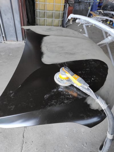 This image depicts a car hood being sanded in what appears to be a workshop or garage. A power sander with a yellow and gray body is positioned on the dark surface of the hood, engaged in sanding a lighter, matte area. The hood itself is angled, resting on an unseen surface. In the background, there are large industrial containers, shelving units, and other car parts, including what looks like a white bumper on a stand. The floor is concrete. There are no people visible in the image. The lighting suggests an indoor setting, possibly with ambient light and artificial illumination. The overall scene conveys a sense of automotive repair or preparation work.