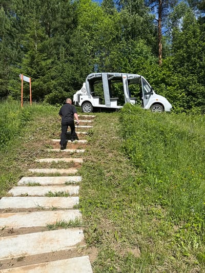 An elderly man wearing a black polo shirt and black trousers is walking up a set of concrete steps that ascend a grassy hill. He is facing away from the camera. At the top of the hill, to the right of the man, is a white, open-sided vehicle, resembling a golf cart or small shuttle bus, with a logo on its side. The vehicle appears to be stationary. The background consists of dense green trees and foliage under a bright, partly cloudy sky. A small signpost with orange poles and a white rectangular sign is visible to the left of the steps. The scene is outdoors and appears to be a park or natural area, likely in or near the village of Nechkino, Russia.