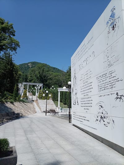 The image shows a view of the monument "Taman Divizion" in the city of Zheleznovodsk, Russia. The monument is a large white wall with bas-reliefs and inscriptions. The wall is decorated with a drawing of a man on horseback and several lines of text. In the background, there is a lush green mountain and a clear blue sky. The foreground shows a paved walkway lined with benches and lampposts. The overall scene is peaceful and serene, with a strong sense of history and culture.