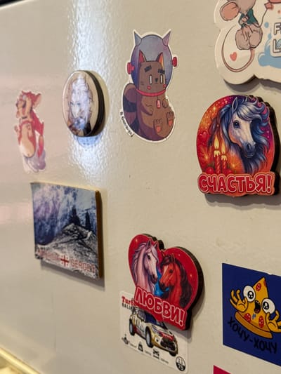 The image shows a close-up of a refrigerator door adorned with various stickers and magnets. The items include cartoonish animal figures, a depiction of horses with text in Russian, a magnet with a landscape scene and text that appears to say "GEORGIA GERGETI," and a sticker of a startled pizza slice with Russian text. The refrigerator door is a neutral, off-white color, and the lighting suggests an indoor environment. There is no discernible context to indicate this is in Яромінскі сельскі Савет, Belarus.