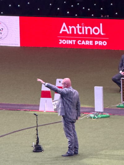 An indoor arena scene on an artificial grass surface, likely at the National Exhibition Centre (NEC) in Marston Green, Metropolitan Borough of Solihull, United Kingdom. A bald man with a beard and glasses, wearing a grey suit, stands with his back to the viewer, extending his right arm forward and slightly upward in a gesture. A black microphone stand with a circular base is positioned on the grass in the foreground. The background features a large LED screen displaying "Antinol. JOINT CARE PRO" on a prominent red panel. To the left of this, a white panel with partial text and a circular graphic is visible. White and red striped pylons, one partially displaying "Crufts," are situated further back on the grass. To the right of the gesturing man, a white rectangular column and green equipment bases with associated cabling are present. A second individual, wearing a dark suit, is partially visible seated on the far right edge of the frame.