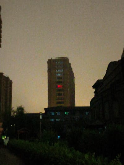 The image depicts an outdoor urban scene, likely at dusk or night, in Jiguan District, China. The focal point is a tall, multi-story building with a distinct red light visible in one of its upper windows. To the left, another, shorter building is partially visible, also appearing to be an apartment complex. In the foreground, dense, dark foliage forms a significant portion of the lower half of the image, obscuring the ground level. A streetlight with a visible lamp post stands amidst the greenery. The sky is overcast with a muted, yellowish-gray hue. The overall image is grainy, suggesting low light conditions or a lower resolution capture. No individuals are clearly discernible in the scene.