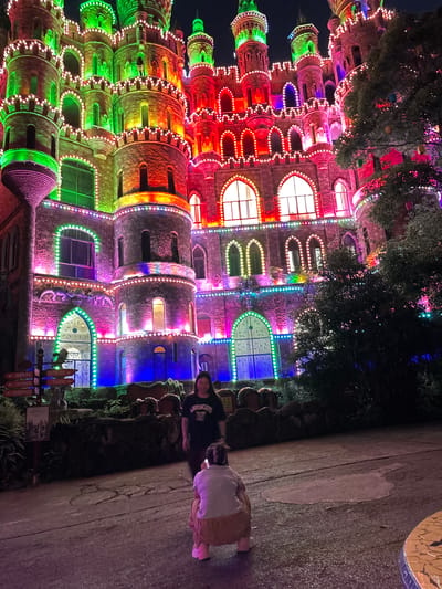 The image captures a view of Lejoy Town in Baqiao, China, at night. The architectural structure is brightly lit with a rainbow of colored lights, emphasizing its castle-like design with multiple turrets and arched windows. In the foreground, two figures, possibly tourists, are visible along a paved pathway. One is wearing dark clothing, while the other is wearing a light-colored top and khaki bottoms. The path is lined with decorative elements, including what appear to be sculpted barrels or similar objects. Signage is present, likely providing information about the location or directions within the town. Trees and vegetation frame the building, adding to the overall ambiance.