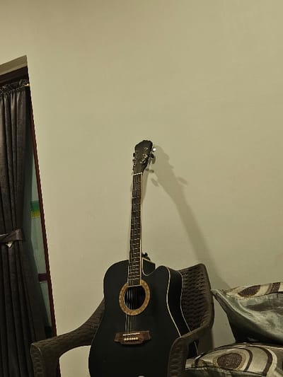 The image captures an indoor scene in the city of Padajaya, Indonesia. A black acoustic guitar with a natural wood rosette and bridge, featuring six strings and a cutaway body, stands upright, resting against a dark brown, woven-style chair. To the left, a dark brown curtain covers part of a window or glass door, revealing narrow vertical segments of colored light, primarily green and a reddish-purple hue, along the edge. A light-colored wall serves as the primary background, displaying a prominent shadow cast by the guitar to its right. To the right of the chair, two patterned cushions, one partially visible, are present, showing a light blue-grey fabric with dark circular designs. No individuals are depicted, and no actions or interactions are occurring.