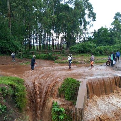 The image depicts a flooded area in Keyian ward, Kenya. A group of people are wading through the water, which is flowing over a small dam or spillway. A boy stands on higher ground to the left, while three individuals cross the watercourse further up. One carries a bundle. Next to them, a woman walks in the water. To the right, two men stand near a red motorcycle. The background features a line of trees and vegetation. The water is heavily discolored, appearing muddy.
