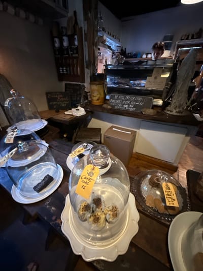 The image shows a collection of bakery items and goods in a shop. It is likely located in Hout Bay, South Africa based on the pricing signs. A sign says "WE STILL PREFER CASH PURCHASES UNDER REO add R2 AMEX CARD ADD 3% ALL RIGHTS RESERVED" with REO referring to ZAR currency. Another sign advertises banana bread.
Visible items include:
* Cookies displayed on a tray with a cover.
* Carrot cake under a glass dome.
* Other cakes in glass containers.
* A blackboard indicating "Banana Bread".
* A wine rack.
The lighting indicates an indoor setting. The overall impression is of a small bakery or deli-style environment.