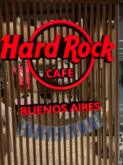 The image shows a neon red "Hard Rock Cafe" sign and the words "Buenos Aires" in red neon letters. The sign is situated in front of a wooden slatted wall, and a string of small Argentinian flags hangs to the right of the "Buenos Aires" text. The flags are light blue and white striped with a sun emblem in the center. The setting appears to be indoors, possibly a commercial establishment, given the presence of merchandise on shelves behind the sign. Some people can be seen in the background, reflected in a glass surface above the wooden slats, suggesting this is a public space. The lighting is artificial, typical of an indoor environment. The overall impression is that of a branded establishment in Argentina.