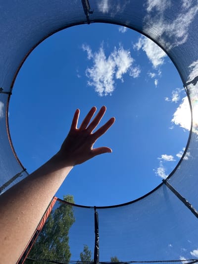 An arm and hand are reaching upwards towards a bright blue sky filled with scattered white clouds. The viewpoint is from inside a trampoline, looking out through the safety net. The edge of the trampoline's dark netting curves around the frame, with hints of orange padding visible underneath. Trees can be seen in the lower left and right corners of the frame, suggesting an outdoor setting, likely a backyard in Novozhilovo, Russia. The lighting indicates a sunny day.