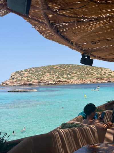 This image shows a view of a beach with clear turquoise water and a rocky, vegetated cliff face. The scene is viewed from under a thatched roof structure, with part of the roof and a speaker visible in the foreground. In the water, there are several people swimming, a yellow floating object, and a small white boat. The sun is shining brightly, suggesting a warm, clear day.

The perspective of the photo is tilted, making it appear as if the beach and sea are on a side plane. In the right side of the image, a person is sitting on a bench-like structure, looking at a phone. Their body is partially in shadow cast by the thatched roof, creating a striped pattern on the bench. The overall atmosphere appears relaxed and leisurely, typical of a beach vacation. The location is identified as Sant Josep de sa Talaia, Spain, which is known for its beautiful beaches. There is no visible text in the image.