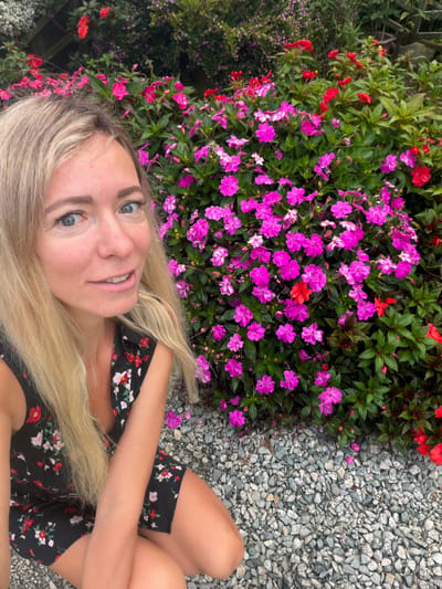 In Xuan Truong - Da Lat, Vietnam, a blonde woman with light blue eyes and fair skin is captured in a selfie. She is wearing a black dress with a floral pattern featuring red and white flowers. The woman is positioned in the foreground, with her face partially visible and looking towards the camera. Behind her, a vibrant display of pink and red impatiens flowers fills the background, intermingled with lush green foliage. The ground is covered in small, grey gravel. The image appears to be taken outdoors, with natural lighting.
