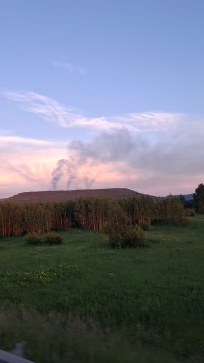 The image depicts a rural landscape under a dusky sky, with the sun setting or rising and casting a soft, colorful glow. The scene is dominated by a sloping hill covered in a dense forest of deciduous trees, their foliage a mixture of green and autumnal hues. The ground leading up to the trees is a verdant meadow dotted with small yellow wildflowers.
The sky transitions from a soft blue on the left to streaks of pink and orange clouds on the right, suggesting the twilight hour. Wisps of dark smoke rise from behind the trees on the left side of the image, indicating a potential controlled burn or a distant fire. The overall atmosphere is serene and natural, with a touch of mystery due to the smoke.
The perspective is from a slightly elevated viewpoint, possibly from a road or a higher point of land, looking across the meadow and up the forested slope. The image is oriented vertically, with the sky occupying the left half and the land the right half. No people or man-made objects are clearly visible, emphasizing the natural setting. The lighting suggests either early morning or late evening, with the light being soft and diffused. The location context of Mazul'skiy, Russia, suggests a temperate climate where such autumnal colors and potential controlled burns might be common.
