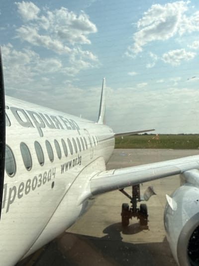 An aerial view from inside an airplane shows clouds in the sky and the side of the aircraft. The plane's text reads "air.bg" and "prevozov". The landing gear is visible on the tarmac. The location is Hostivice, Czechia.