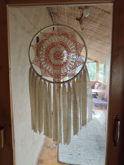 A handmade dreamcatcher, with an orange and white crocheted circular web, hangs in a doorway. It is adorned with numerous white crocheted fringe tassels. The dreamcatcher is positioned in front of a glass door, reflecting an interior space with a light-colored rug and wooden floorboards. Beyond the glass, the interior appears to be a sunroom or porch with large windows offering a view of green foliage. A wooden table and seating are visible within this enclosed space. The exterior wall to the left of the dreamcatcher is textured and off-white. The ceiling above the dreamcatcher features a woven bamboo-like material and a simple light fixture. The overall scene suggests a rustic, possibly bohemian, aesthetic within a residential setting in the mountain village of Prini, India.