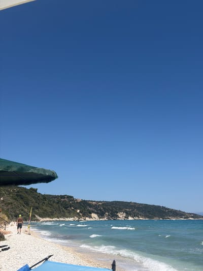 The image depicts a vibrant coastal scene with a clear blue sky dominating the left side. On the right, a pebbled beach stretches towards a shimmering turquoise sea, where gentle waves are breaking. Lush green vegetation covers a sloping hillside that forms the backdrop to the beach.

In the foreground on the beach, a person is seen walking away from the camera. They are wearing black shorts and a yellow cap, with a backpack visible on their back. Further down the beach, two other figures are walking. A yellow windsurfing sail with a stand is propped up on the beach near the water's edge. Two blue umbrellas are visible, one on the far right and another partially obscured in the bottom left, providing shade on the sunny beach.

The scene appears to be set during the daytime, with bright sunlight illuminating the landscape. The weather is clear and sunny, ideal for a day at the beach. The location cues suggest a Mediterranean setting, consistent with the provided location context of Elaties Municipal Unit, Greece. The overall mood is one of relaxation and leisure. No visible text is present in the image.