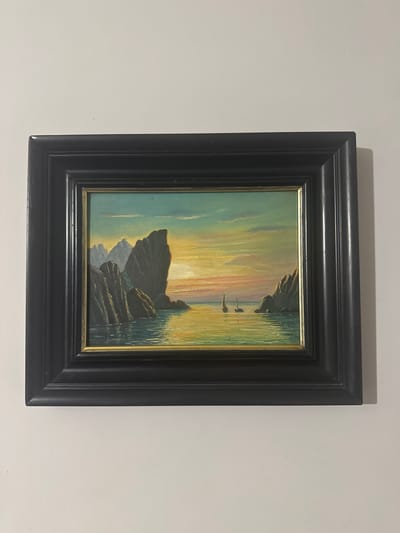 The image displays a framed painting of a seascape with sailboats at sunset. The painting is hung on a white wall. The seascape features jagged rock formations jutting out of the water, with a colorful sky transitioning from yellow and orange near the horizon to blue and green above. Two sailboats with white sails are visible on the calm water, reflecting the warm hues of the sunset. The painting is enclosed in a dark, wide frame with a textured surface. The overall scene is peaceful and idyllic. There is no information in the image to suggest the location in the city of Novosiltsi, Ukraine.