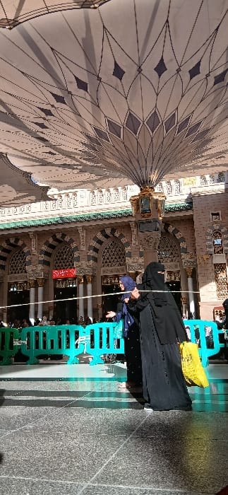 The image captures an outdoor courtyard scene at the Masjid al-Nabawi (Prophet's Mosque) in Medina, Saudi Arabia. A large, ornate retractable umbrella, beige with dark geometric patterns, dominates the upper portion of the frame, casting shadows. In the mid-ground, two women are positioned near a line of light green plastic crowd control barriers and a white rope. The woman on the right wears a black niqab and abaya, carrying a bright yellow plastic bag in her left hand while her right hand appears to grasp the white rope. The woman on the left wears a dark blue hijab and abaya, with her face visible, and extends her left arm in a gesturing motion. The background reveals the ornate architecture of the mosque, including columns with gold-colored capitals, arched sections with decorative grilles, and a prominent green-tiled roofline. A red sign with white Arabic text is visible on a background wall. The ground consists of light grey speckled paving stones, illuminated by direct sunlight which creates distinct shadows.