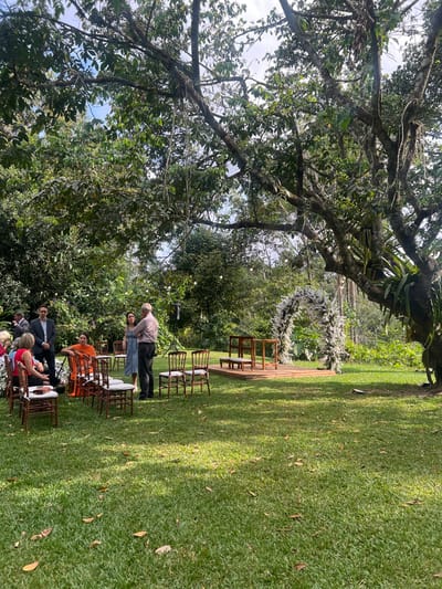 The image depicts an outdoor event venue in Camaragibe, Brazil, characterized by a manicured grass lawn bordered by dense tropical vegetation under a partly cloudy sky. A large, mature tree with thick branches and epiphytes anchors the right side of the frame. A ceremonial area is prepared with a raised wooden platform, featuring two unadorned wooden structures, possibly altars or stands, and a prominent white floral arch composed of white flowers and green foliage positioned behind them. Brown wooden chairs with light cushions are arranged in rows on the lawn, facing the platform; some are occupied. Approximately ten individuals are present. On the left, a man in a dark suit jacket stands near seated guests, including a woman in a red top. Centrally, an older man in a light-colored polo shirt and dark trousers is conversing with a woman in a light blue dress. Another woman in an orange shawl or dress is seated adjacent. Decorative string lights are suspended among the tree branches, and a black speaker on a stand is positioned to the left of the platform. The scene indicates a social gathering or ceremony in a natural, landscaped setting.