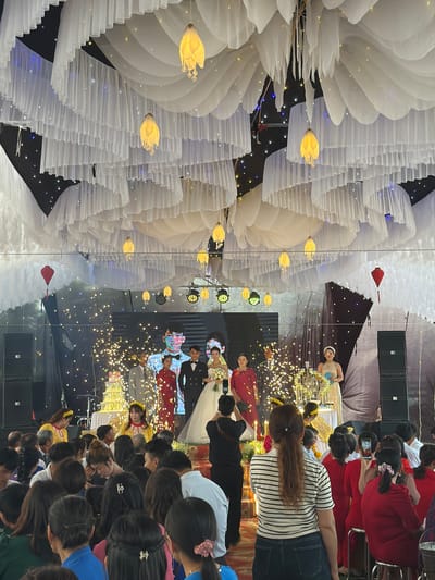 The image depicts an indoor wedding event, likely in Bình An, Vietnam. The venue is decorated with large white floral arrangements and strings of lights. A stage is visible with several individuals, possibly the wedding party, dressed in formal attire. Attendees are observing the event; some are taking pictures. Sound equipment is present, indicating audio amplification. The environment suggests a festive and celebratory atmosphere.