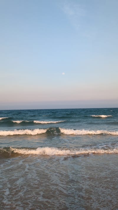 The image depicts a beach scene in Unknown, Vietnam. The left side of the image features a light blue sky with a small, circular object resembling the moon. To the right of the sky is the ocean, with visible waves crashing onto the shore. The water color transitions from a deep blue further out to a lighter, sandy hue near the beach. The waves are foamy and white-capped. The image is oriented in a portrait style, with the horizon line running vertically down the center. There are no people visible in the image.