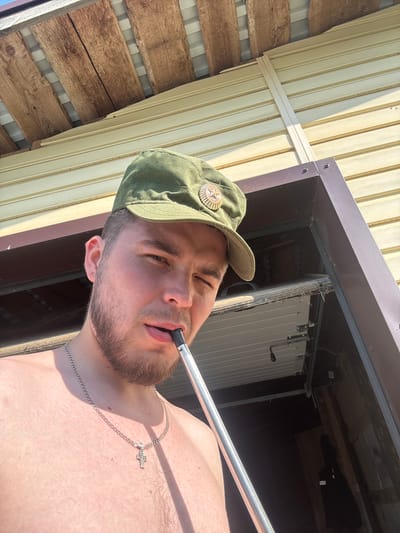 A young man, wearing a green military-style cap with a star emblem, is taking a selfie outdoors. He has a beard and is wearing a silver chain with a cross pendant. He is holding a hookah mouthpiece to his lips, suggesting he is smoking. The background shows the exterior of a building with light-colored horizontal siding, and above that, wooden planks are visible, possibly part of a roof structure. The lighting suggests it is a sunny day. The location is identified as Borodulino, Russia.