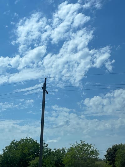 The media file shows a view of the sky with scattered clouds, a utility pole, and power lines. The sky is a bright blue, suggesting daytime. The clouds are white and wispy, indicating fair weather. Green foliage from trees is visible on the right side of the frame, hinting at a natural or semi-rural environment. The utility pole is dark and positioned diagonally across the frame, with several power lines extending from it. There are no people or animals visible in the image. The overall impression is one of a serene, outdoor scene under a clear sky. The location context provided is Donskoe, Russia. There is no visible text in the image.
