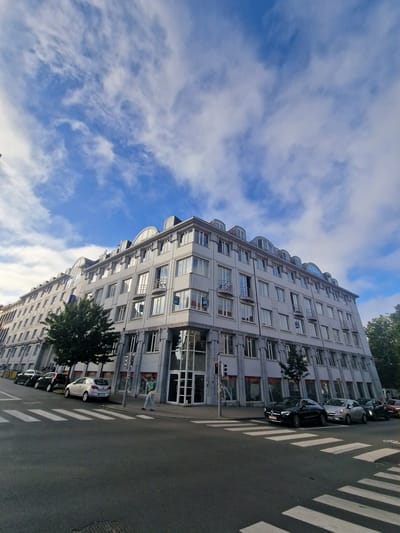 This is a low-angle, wide-shot photograph of a light-colored, multi-story building with a curved roofline, situated at a street corner in Saint-Gilles - Sint-Gillis, Belgium. The building features numerous windows, some with balconies, and a modern architectural style with a stone-like facade on the lower floors and lighter material on the upper floors. A few trees are visible alongside the building, and several cars are parked along the street. In the foreground, a pedestrian crossing is marked on the asphalt road. A single person, wearing a green top and blue jeans, is walking on the sidewalk near the building, facing away from the camera. The sky above is a bright blue with scattered white clouds. The scene appears to be a typical urban street view.
