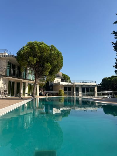 The image depicts a bright, sunny day in Brca, Montenegro. In the foreground, a large, turquoise swimming pool with tiled bottom reflects the surrounding environment. The pool is bordered by a light-colored deck made of wood planks.
To the left of the pool, a modern building with multiple levels is visible. The building features light-colored stone walls, large glass windows, and balconies with metal railings. A mature pine tree stands prominently in front of this building. Two outdoor chairs and a small table are placed on the deck near the pool.
Further back, another section of the building complex is visible, also with balconies and glass windows, set against a backdrop of lush green trees and a clear blue sky. The overall scene conveys a sense of luxury and relaxation, typical of a resort or private villa. There are no people or animals visible in the image.