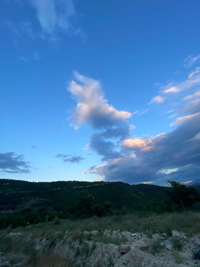 The image depicts a landscape with rolling hills covered in dark green foliage under a bright blue sky with scattered clouds. The foreground shows a rough, rocky terrain with dry grass. The scene appears to be a rural or natural setting, possibly on the outskirts of the city of Ablanitsa, Bulgaria. No people or specific objects are visible, and no actions are taking place. The overall impression is of a serene, natural environment during daylight hours.