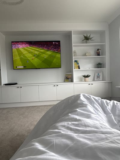 The image shows a scene inside a room with a white wall-mounted shelving unit. The unit has open shelves at the top displaying decorative items, including potted plants, a toucan figurine, and cards/small books. Beneath the shelves, there's a television displaying a football game. The lower part of the shelving unit consists of closed cabinets with silver round doorknobs. A white bed with rumpled white bedding is partially visible in the foreground to the right. The room appears to be in a private residence in Upper Borth, United Kingdom. The OCR text visible on the television screen indicates "LIV 11 CHE 03:06" and "RTS," while a card on the shelf reads "IT'S NOT SCI-FI IT'S WIFI I."