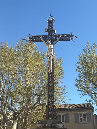 A low-angle shot depicts a sculpture of Jesus Christ crucified on an ornate iron cross, suspended in front of a clear blue sky and foliage. The building on the right is part of the “Musee Frederic Mistral” in Maillane, France. The structure of the cross is elaborately decorated, with spiked finials on top and bottom, as well as scrolled accents. The sculpture of Jesus is positioned horizontally on the cross. The backdrop is a mix of blue sky and trees with light green foliage. A building with two windows and shutters is seen on the right side of the composition.