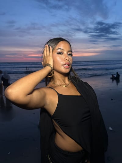 The image displays a twilight beach scene in Kerobokan Kelod, Indonesia. A female subject with medium-to-dark skin stands prominently in the foreground, positioned slightly right of center, looking directly at the viewer. She has dark hair, styled with visible makeup including winged eyeliner and glossy lips. She wears a black sleeveless top with a side cut-out revealing her waist, and a black draped garment over her shoulders. A gold-colored necklace with a circular pendant is visible on her neck, and multiple gold-colored bangles adorn her left wrist. Her left hand is raised to the side of her head, fingers splayed. The background features a wet, dark sand beach transitioning into ocean waves. The sky exhibits a gradient of dark blue, purple, pink, and orange, indicative of twilight, with scattered dark clouds. Several blurred, dark silhouettes of individuals are visible in the midground: one figure stands to the far left, and two distinct figures are positioned near the waterline on the right, with an additional faint figure further out in the ocean.