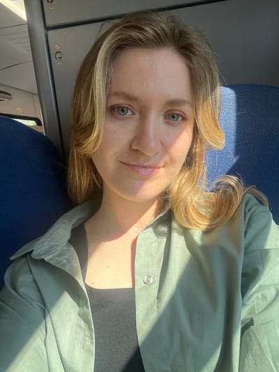 A young, fair-skinned woman with blonde hair is in an indoor setting, possibly on a train or bus. She is wearing a light green button-down shirt over a black top and sitting on a blue seat. Sunlight streams in, casting shadows across her face and clothing. She is looking at the camera, appearing relaxed. The background shows parts of the train interior including an overhead compartment and window. The location is not discernable from the image.