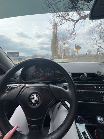 The image displays the interior of a BMW vehicle, specifically from the driver's perspective, looking forward onto a road in Piperkov Chiflik, Bulgaria. The sky is overcast with a dense layer of clouds.

Inside the vehicle, a black leather steering wheel with blue-green contrast stitching and a central BMW logo dominates the lower half of the frame. The driver's left hand, adorned with long pink acrylic nails, is visible holding the bottom left of the steering wheel. The driver's legs, clad in white trousers or sweatpants with a visible dark stripe and text (partially reading "WMES Desserts"), are positioned below the steering wheel.

The dashboard features an instrument cluster indicating a speed of 0 km/h, an engine idle, and a digital display showing "19:23" (time) and "18.2 °C" (temperature), along with a trip reading around "482" kilometers. To the right, the center console includes a radio displaying "FM 97.2," and a magnetic phone mount is affixed to the right-side air vent. A blue-lit electronic device is plugged into a 12V socket near the manual gear shifter, which features a knob with M-stripe accents.

Through the windshield, a paved road extends into the distance, with a clear intersection visible ahead. The environment is characterized by numerous bare deciduous trees, suggesting a non-summer season. A very tall, slender, bare tree, consistent with a poplar, stands prominently on the left side of the road. A large, gnarled bare tree branch extends over the top right of the windshield.

Essential road signage includes a prominent yellow diamond-shaped "priority road" sign on the right side of the road, appearing slightly worn or bent. Another smaller red and white circular sign is partially visible on the left, behind the tall poplar tree. Power lines and utility poles traverse the scene. Two dark-colored vehicles are visible on the road in the distance, while a larger, possibly industrial, vehicle is partially discernible on the far left. Distant hills or mountains are present on the horizon beneath the cloudy sky. A rustic fence line runs along the right side of the road.