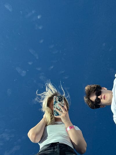 The image shows a reflection of two people against a blue surface. A person with sunglasses is positioned near the top, appearing upside down. A person holding a phone with a floral case is positioned towards the lower right, taking a picture, and appears sideways. The blue surface has some smudges and imperfections on it.