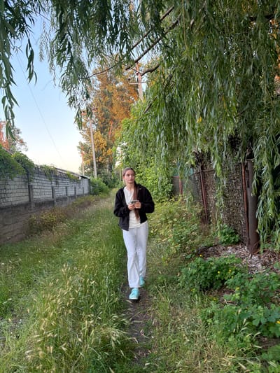 A young woman walks along a grassy path in Obukhivka, Ukraine. She is wearing a black zip-up jacket over a white t-shirt and white sweatpants. Her light blue sneakers are visible as she walks. She holds a white smartphone in her right hand. The path is bordered on the left by a long, low brick wall topped with greenery. On the right, the path is shaded by the drooping branches of a large tree, and a rusty metal fence is visible. Tall grasses and wild plants grow along the path and around the fence. In the background, a telephone pole and trees are visible against a clear, pale blue sky. The light suggests it is either early morning or late afternoon.