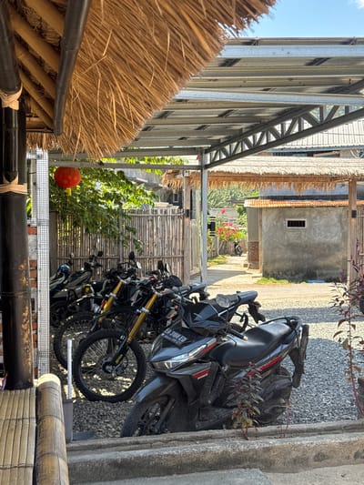 The image depicts a motorcycle parking area outside a building with a thatched roof and a metal awning in Kertasari, Indonesia. Several black motorcycles are parked on a gravel surface. A red lantern hangs from a bamboo pole. In the background, a building with a grey concrete exterior and an orange tiled roof is visible, along with a wooden fence and greenery. The scene appears to be outdoors in daylight, with a clear blue sky overhead. No people are present in the image.