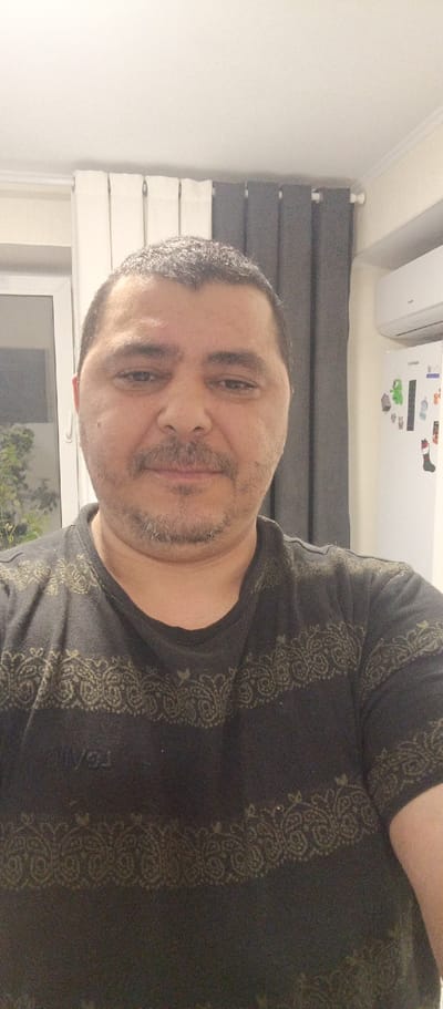 A middle-aged man with short, grey-flecked hair and a light beard is shown in a selfie taken indoors. He is wearing a dark t-shirt with a repeating patterned design. The man is positioned in front of a window with white and grey curtains. To his right, a white refrigerator with magnets and an air conditioning unit are visible on a cream-colored wall. The overall scene suggests an indoor domestic setting. The location is not identifiable within the image.
