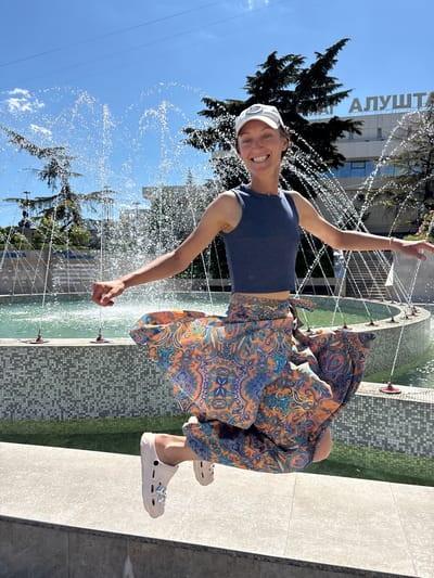 A woman in a blue tank top, a white baseball cap, and a flowing, colorful patterned skirt is captured mid-air as she jumps in front of a fountain in Alushta, Ukraine. She is smiling broadly and her arms are outstretched. The fountain has multiple streams of water spraying upwards, with a tiled basin. In the background, a building with the word "АЛУШТА" (Alushta) written on it is visible, along with trees and a clear blue sky. The woman is wearing light-colored clogs.