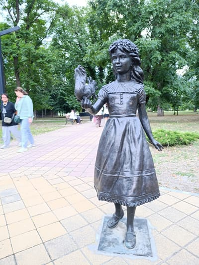 A bronze statue of a young girl stands in a park, holding a rabbit in her right hand. The girl is wearing a dress with a ruffled collar and a wreath of flowers on her head. She stands on a square base. The statue is positioned on a paved walkway in what appears to be a park or garden area. In the background, trees and foliage are visible, and other people are walking on the path or sitting on a bench. The location is identified as Kelermesskaya, Russia.