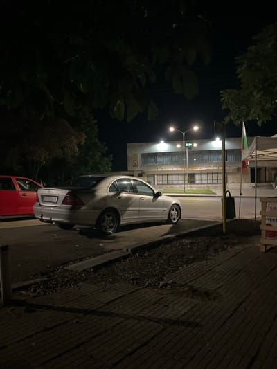 The image depicts a nighttime scene in Krushovitsa, Bulgaria. A silver Mercedes-Benz C-Class sedan is parked on a street next to a red sedan. In the background, a building with illuminated windows stands, and two streetlights illuminate the area. A Bulgarian flag is visible on the right side of the image, attached to a pole. There is also a white canopy structure, possibly a market stall, in the foreground on the right. The ground in the foreground is paved with bricks and shows some areas of gravel and dirt. The overall impression is a quiet street scene at night, with the building and streetlights providing the main illumination.